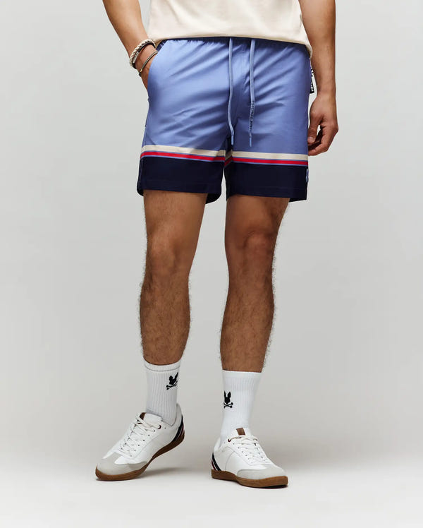 A person wears Psycho Bunny MENS HANKS HYDROCHROMIC SWIM TRUNK - MB1000041 in light purple with navy and white stripes, paired with a white tee, white crew socks with a black logo, white sneakers, and bracelets against a neutral background.