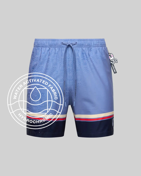 Psycho Bunny MENS HANKS HYDROCHROMIC SWIM TRUNK (MB1000041): Light blue swim shorts with navy accents, red/white hem stripes, drawstring waist, hydrochromic tech revealing "Water-Activated Fabric" graphic, and a cartoon skull tag.