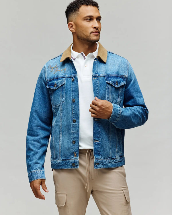 A man stands against a plain background, wearing the Psycho Bunny MENS WADE DENIM TRUCKER JACKET - MO1100015 with a brown corduroy collar over a white polo and beige pants, facing slightly to the side while adjusting his jacket.