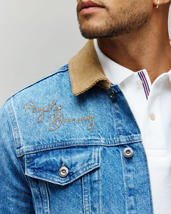 A man wears the Psycho Bunny MENS WADE DENIM TRUCKER JACKET (MO1100015), featuring a blue denim body, tan corduroy collar, and embroidered logo on the chest, layered over a white shirt with a purple-striped placket. His face is partially visible.