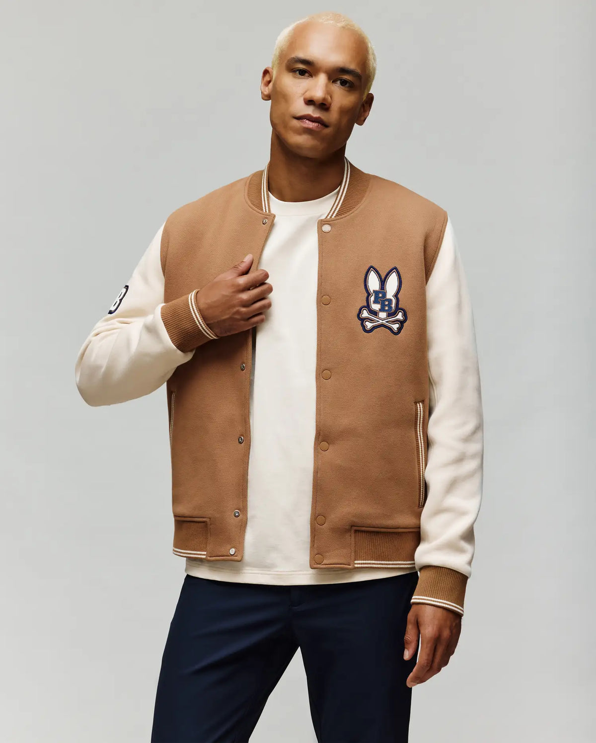 A man with short blond hair models the Psycho Bunny MENS WADE KNIT BOMBER (MO1100020), a tan and white varsity style jacket with an embroidered bunny logo on the chest and striped cuffs, over a white shirt and dark pants.