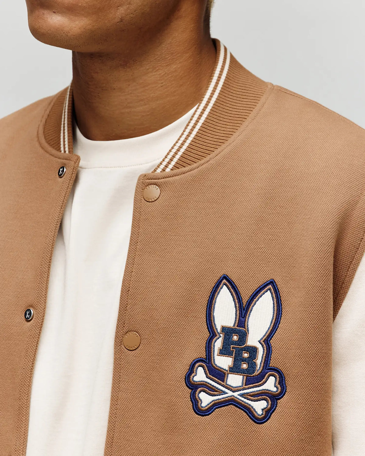 A person wears the Psycho Bunny MENS WADE KNIT BOMBER (MO1100020), a tan varsity-style jacket with white trim, a white shirt, and an embroidered logo of two bunny ears above crossed bones and "PB" lettering.