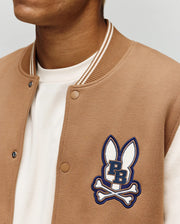 A person wears the Psycho Bunny MENS WADE KNIT BOMBER (MO1100020), a tan varsity-style jacket with white trim, a white shirt, and an embroidered logo of two bunny ears above crossed bones and "PB" lettering.
