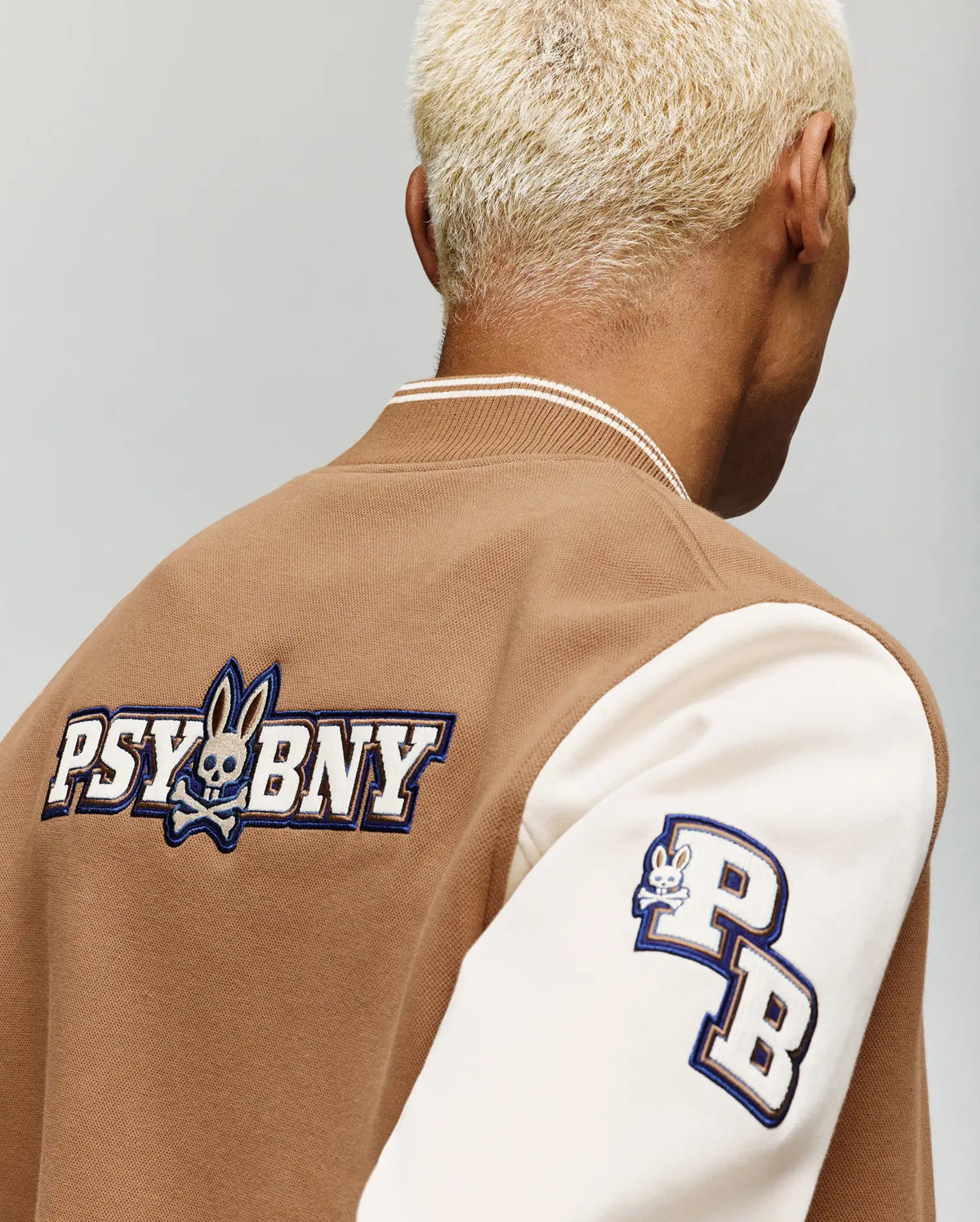 A person with short, bleached blonde hair wears the Psycho Bunny MENS WADE KNIT BOMBER (MO1100020), a tan and white varsity jacket featuring "PSYBUNY" and an embroidered bunny logo on the back, plus "PB" with a bunny logo on the left sleeve.