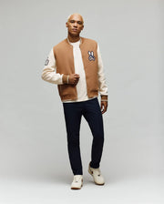 A man with short blond hair models the Psycho Bunny MENS WADE KNIT BOMBER (MO1100020), a tan and cream varsity jacket with rabbit patch, over a cream shirt, dark pants, and white sneakers against a plain light gray background.