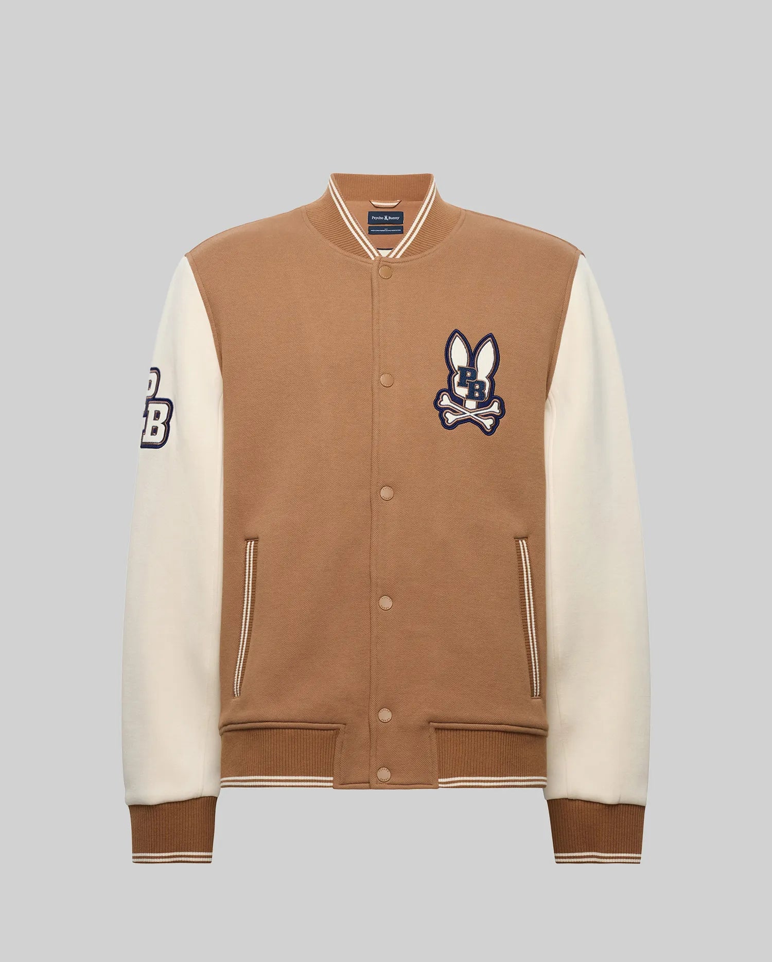 The Psycho Bunny MENS WADE KNIT BOMBER (MO1100020) is a tan and white varsity jacket with a tan body, white sleeves, striped cuffs and hem, snap buttons, and "CB" bunny logo patches on the chest and left sleeve.