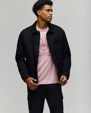 A man stands against a plain background, wearing the Psycho Bunny MENS AVEN KNIT SHACKET (MO1100035) over a light pink T-shirt featuring a stylized bunny graphic, paired with black pants. He looks to the side, one hand adjusting his jacket.