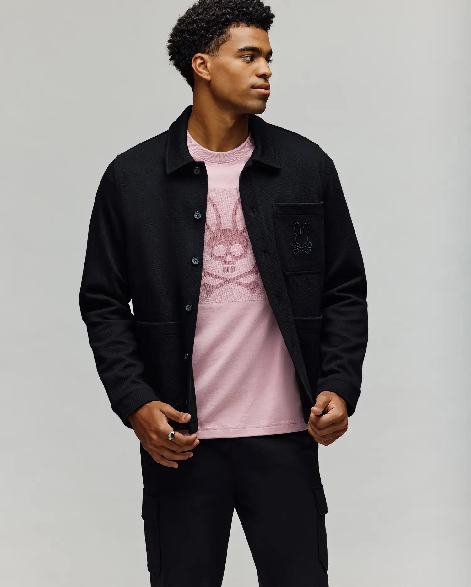 A man stands against a plain background, wearing the Psycho Bunny MENS AVEN KNIT SHACKET (MO1100035) over a light pink T-shirt featuring a stylized bunny graphic, paired with black pants. He looks to the side, one hand adjusting his jacket.