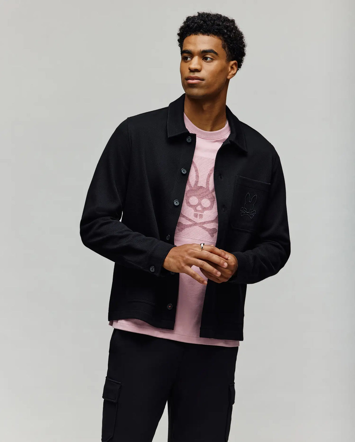 A young man with short curly hair models the Psycho Bunny MENS AVEN KNIT SHACKET (MO1100035) in black over a bunny graphic pink t-shirt and black cargo pants, standing against a plain light gray background—ideal for transitional layering.