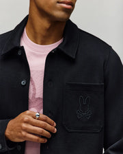 Styled for transitional comfort, a person wears the Psycho Bunny MENS AVEN KNIT SHACKET (MO1100035) with a bunny and crossbones pocket patch over a light pink tee featuring a matching bunny design, accessorized with a silver ring.