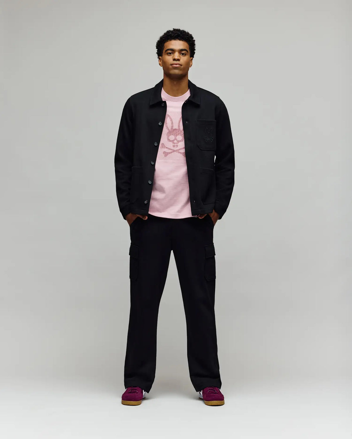 A man faces forward in a Psycho Bunny MENS AVEN KNIT SHACKET (MO1100035), black cargo pants, a light pink graphic tee, and red sneakers, all set against a plain gray backdrop.