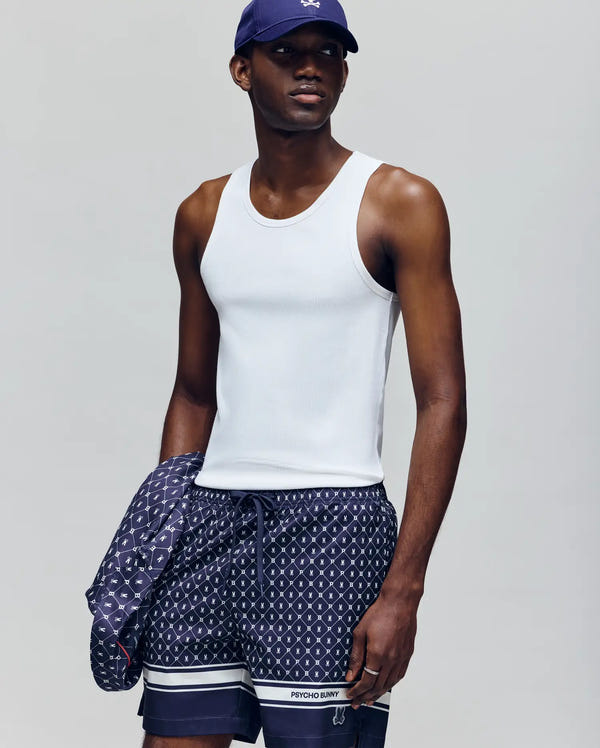 A man wears a Psycho Bunny MENS 2 PACK TANK TOP - MO1400001 in white, navy patterned shorts, and a navy cap, holding a matching jacket. He stands against a plain light gray background, looking slightly to the side.