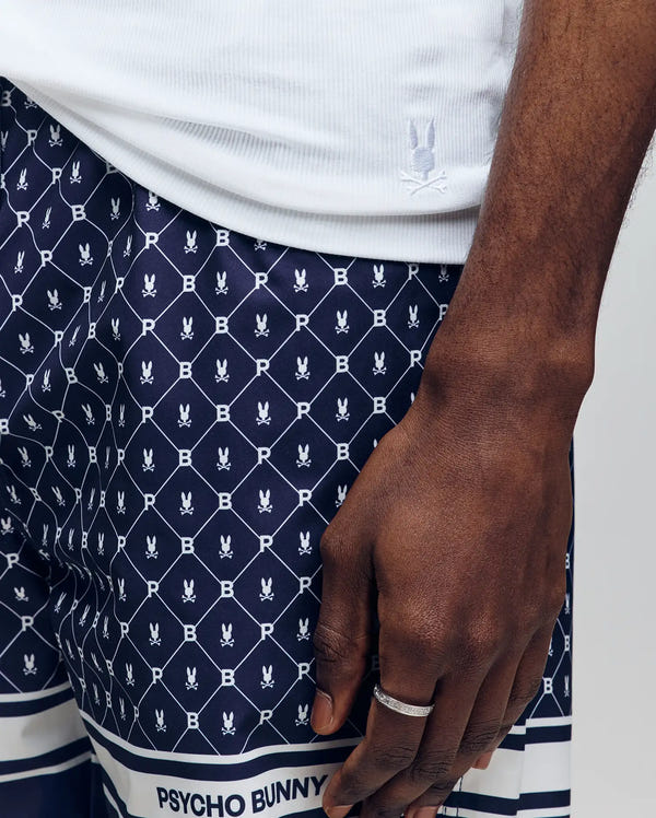 A close-up shows a person wearing the Psycho Bunny MENS 2 PACK TANK TOP - MO1400001 in white, paired with navy shorts featuring a white bunny and "B" print, plus a Psycho Bunny logo patch. They also wear a silver ring.