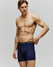A muscular man models the Psycho Bunny MENS 3 PACK BOXER BRIEF - MO1400006, featuring navy blue cotton-modal boxers with a black "Psycho Bunny" waistband. He stands against a plain background, gazing left with one hand on his waistband.