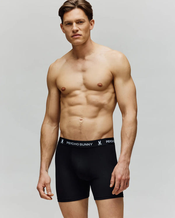 A fit, shirtless man stands against a plain background, wearing Psycho Bunny MENS 3 PACK BOXER BRIEF - MO1400006 in black, crafted from premium cotton-modal for comfort. The waistband features "Psycho Bunny" branding in white text.