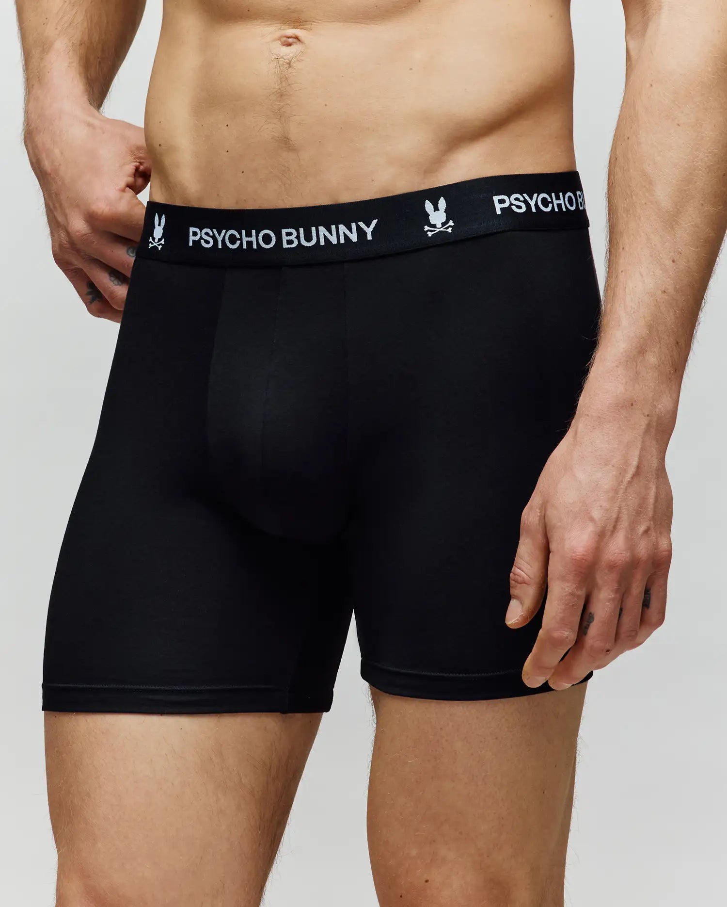 A man wears Psycho Bunny MENS 3 PACK BOXER BRIEF - MO1400006 in black, made from premium cotton-modal with "Psycho Bunny" and bunny logo on the waistband, showing comfort and a close-up of his midsection, shorts, torso, and an arm.