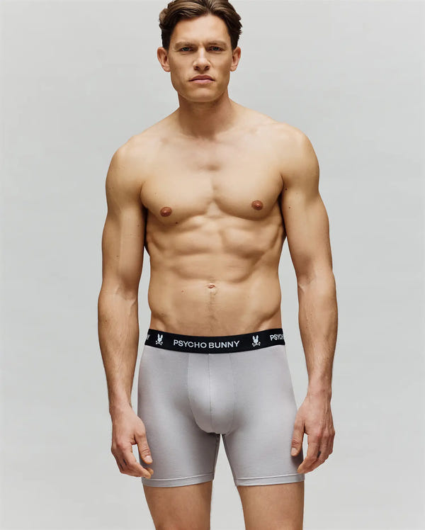 A fit, shirtless man poses against a plain background, wearing Psycho Bunny MENS 3 PACK BOXER BRIEF - MO1400006 in light gray with a soft cotton-modal blend and a black "Psycho Bunny" branded waistband for all-day comfort.