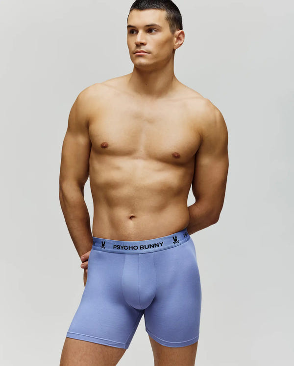 A shirtless man stands against a plain background wearing Psycho Bunny MENS 3 PACK BOXER BRIEF - MO1400008. The light purple modal blend boxers feature "PSYCHO BUNNY" and bunny logos on the waistband, highlighting stylish men's underwear.