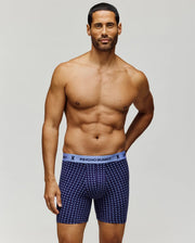 A muscular man with short dark hair stands against a plain background, wearing Psycho Bunny's MENS SINGLE BOXER BRIEF - MO1400010 in patterned navy blue Pima cotton with a light blue "Psycho Bunny" waistband for all-day comfort.
