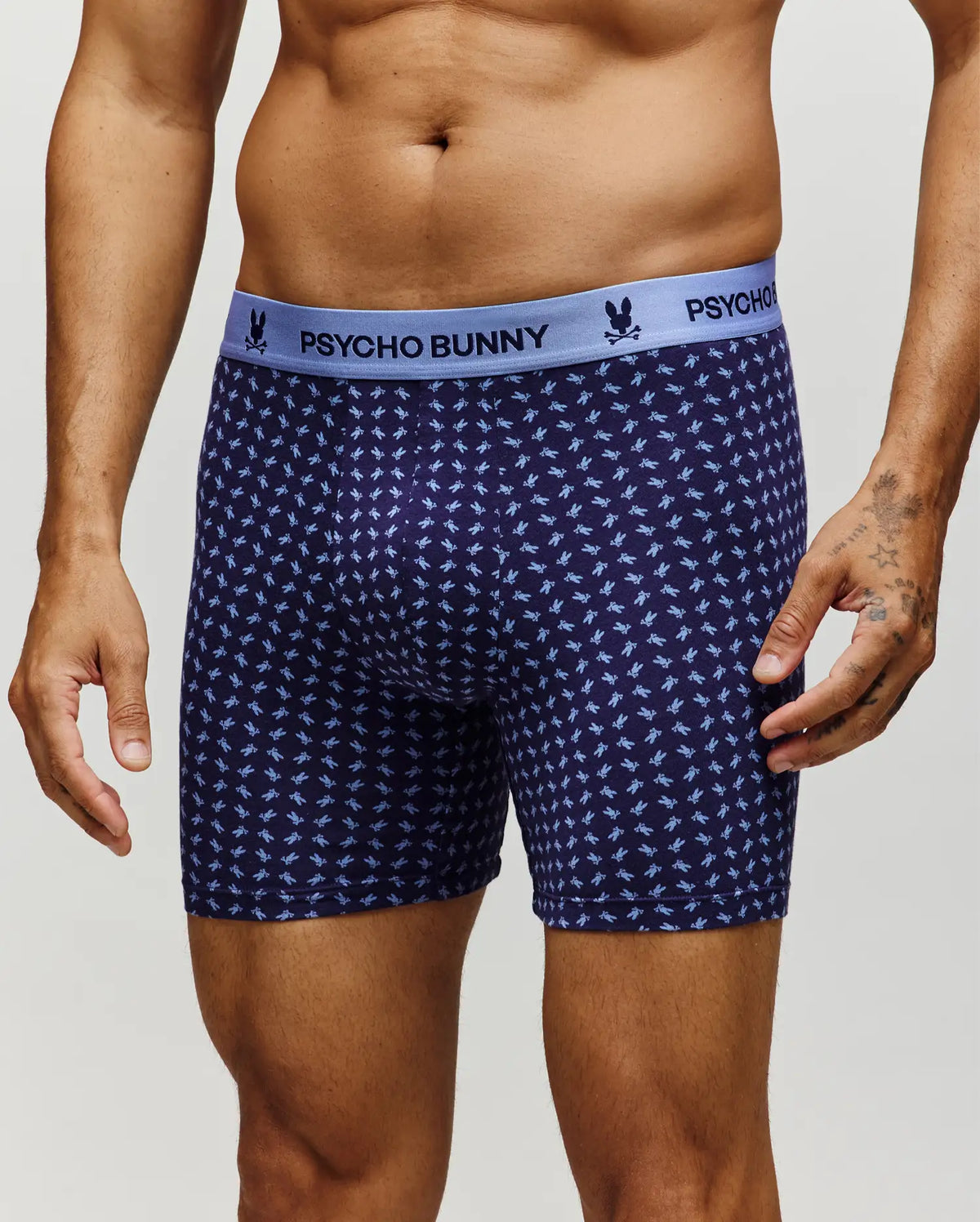 A man models the Psycho Bunny MENS SINGLE BOXER BRIEF - MO1400010 in navy, featuring a light blue waistband with the brand name and a soft Pima cotton fabric adorned with a repeating light blue bunny pattern for all-day comfort.