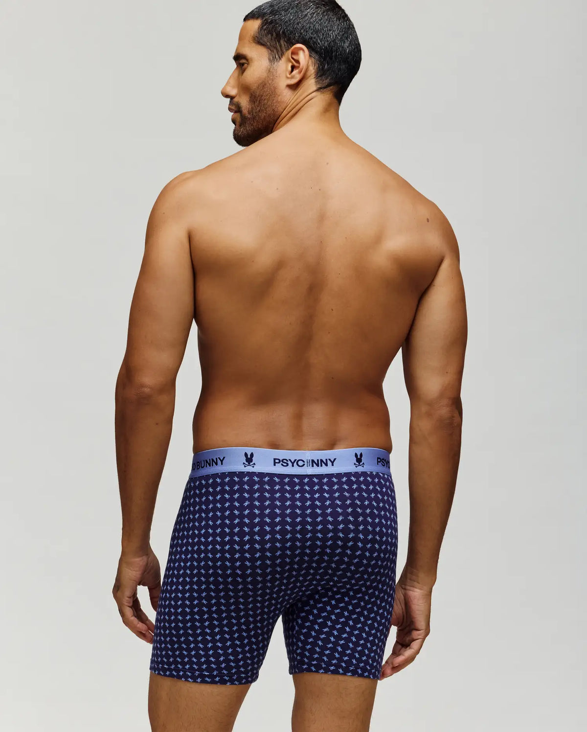 A man with short dark hair stands shirtless, facing away, wearing navy blue Psycho Bunny MENS SINGLE BOXER BRIEF - MO1400010 with a light blue logo waistband and breathable knit Pima cotton for comfort and style.