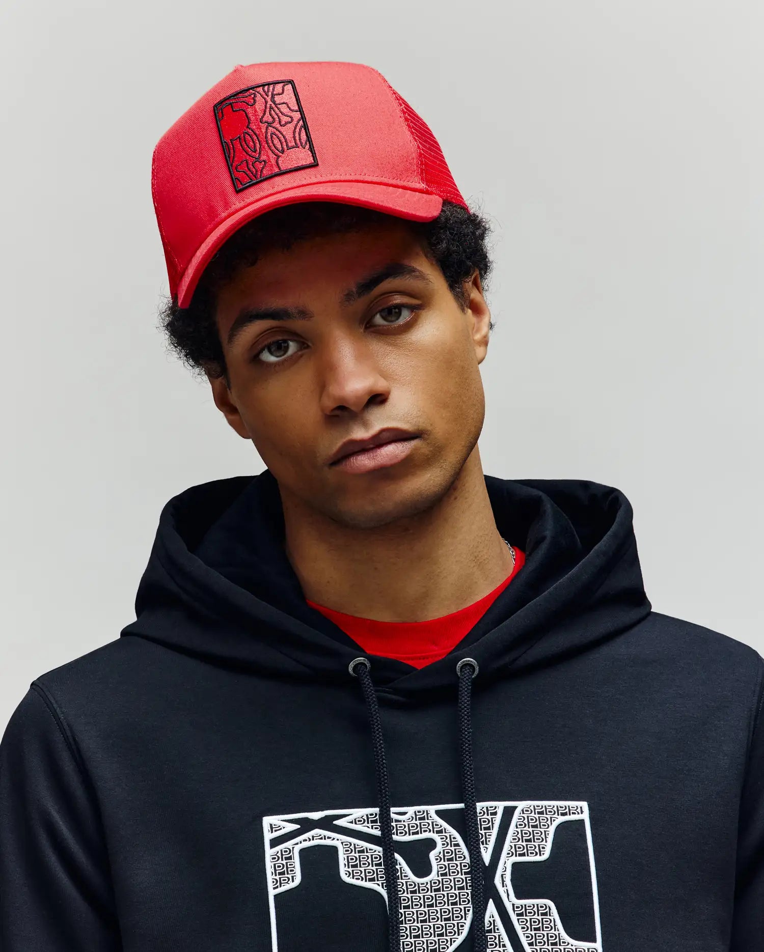 A young person wears the Psycho Bunny MENS GLENN TRUCKER CAP - MO1500004 in red and a black hoodie with Bunnies graphics, standing against a plain light gray background, looking directly at the camera with a neutral expression.