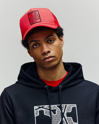 A young person wears the Psycho Bunny MENS GLENN TRUCKER CAP - MO1500004 in red and a black hoodie with Bunnies graphics, standing against a plain light gray background, looking directly at the camera with a neutral expression.