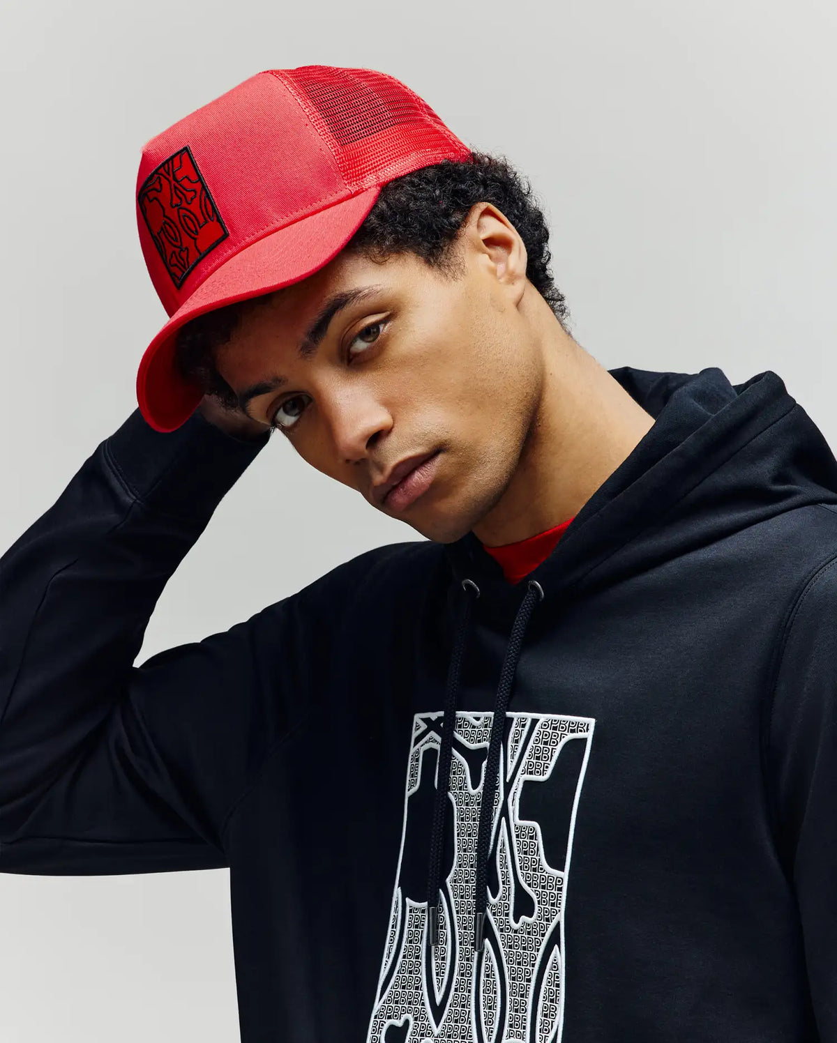 A young man wears the Psycho Bunny MENS GLENN TRUCKER CAP - MO1500004 in red and a black hoodie with a white patterned graphic, looking at the camera with a neutral expression against a plain light background.