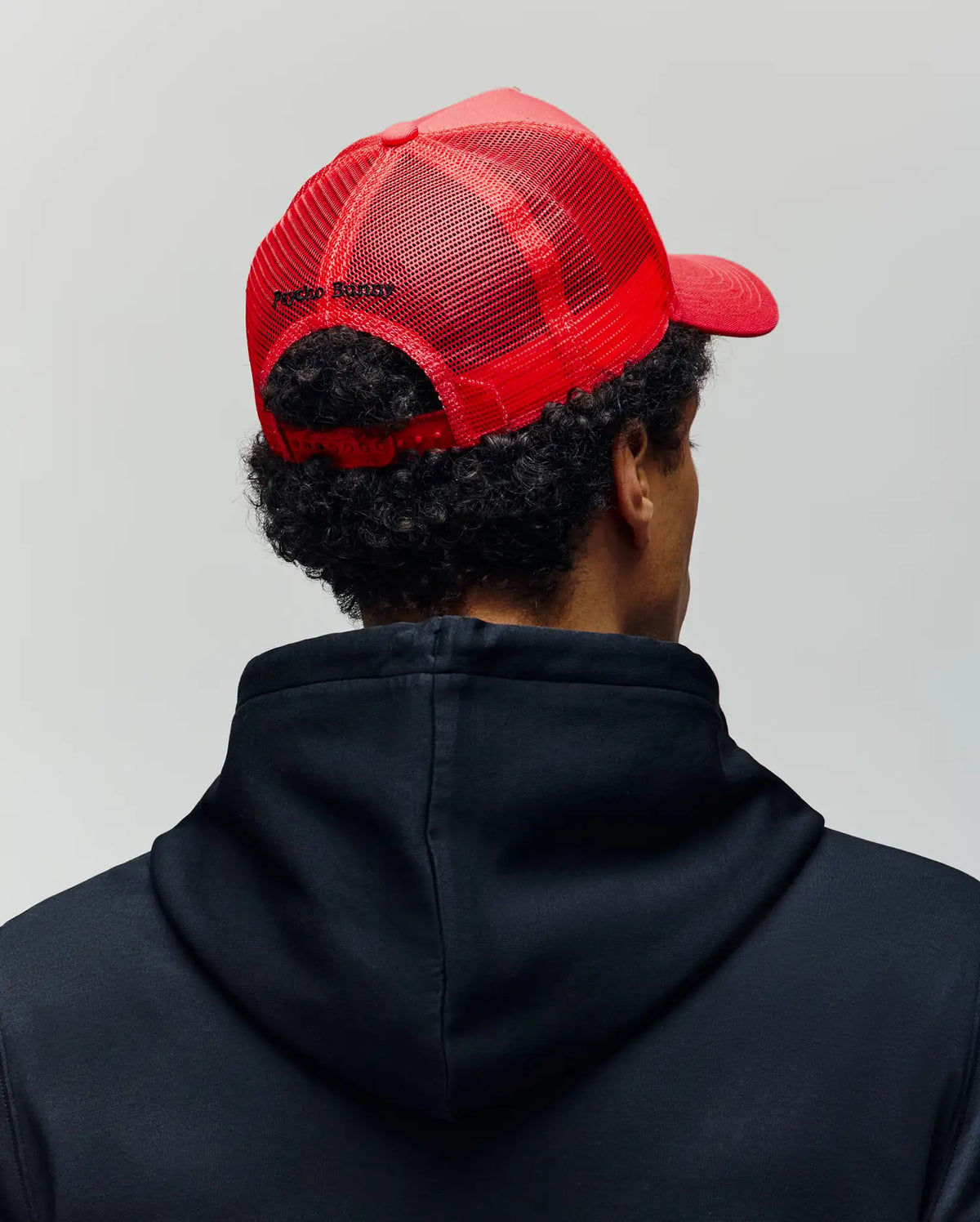 A person is seen from behind against a plain light background, wearing a black hoodie and the Psycho Bunny MENS GLENN TRUCKER CAP - MO1500004, a red mesh trucker cap with an adjustable snapback.