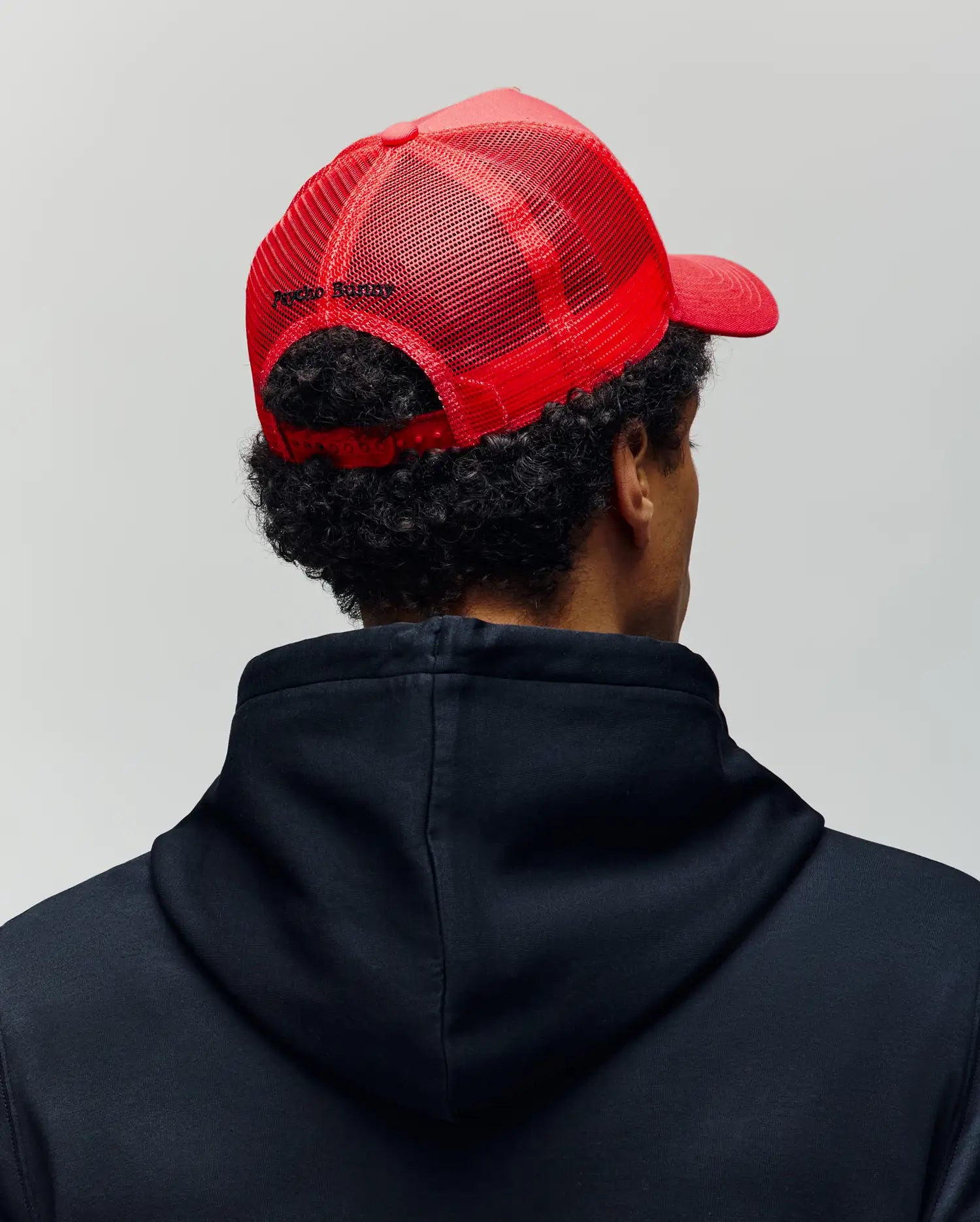 A person is seen from behind against a plain light background, wearing a black hoodie and the Psycho Bunny MENS GLENN TRUCKER CAP - MO1500004, a red mesh trucker cap with an adjustable snapback.