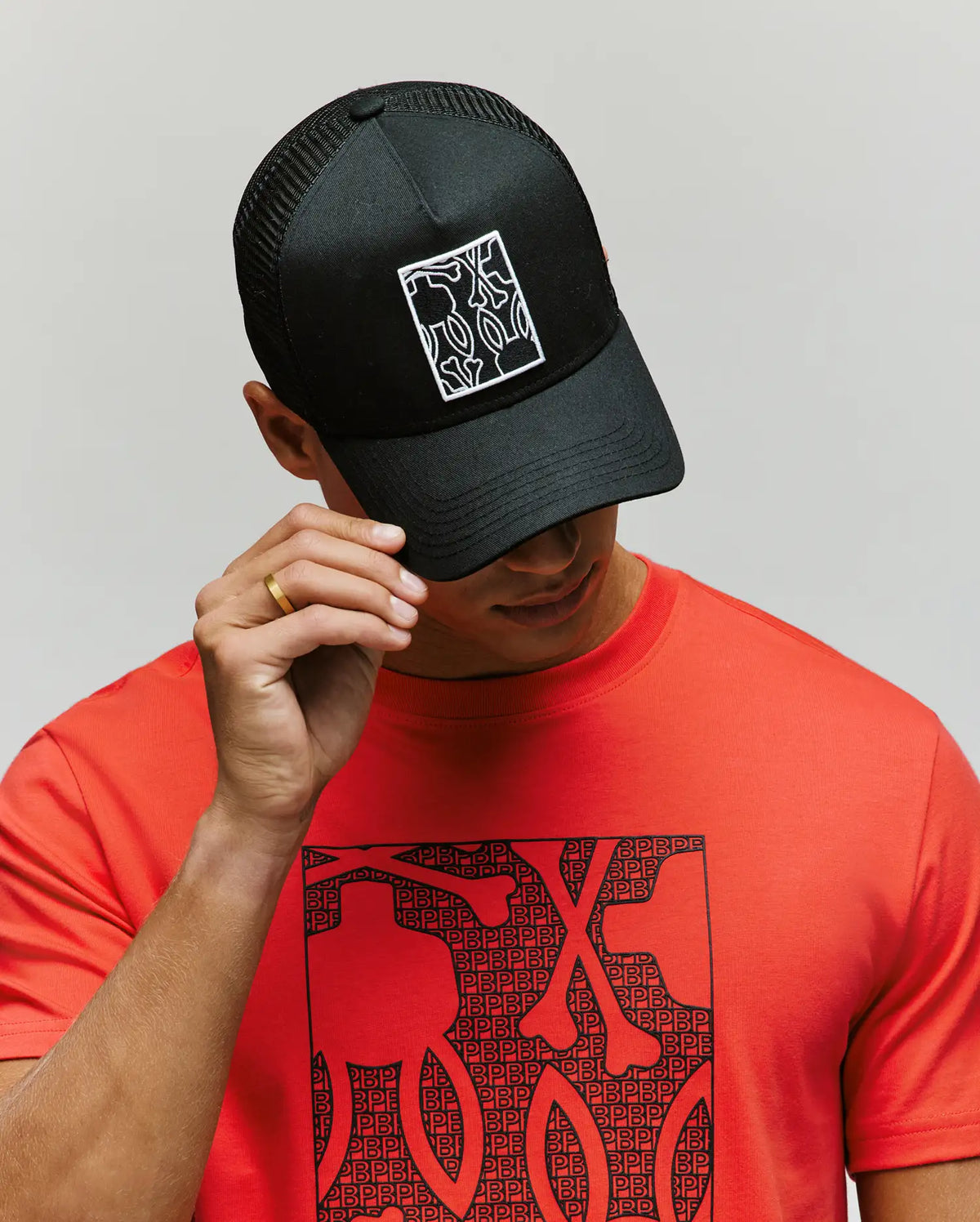 A person wears the Psycho Bunny MENS GLENN TRUCKER CAP - MO1500004, an adjustable black snapback with a white abstract pattern, paired with a red shirt featuring a matching black pattern. The background is plain and light-colored.
