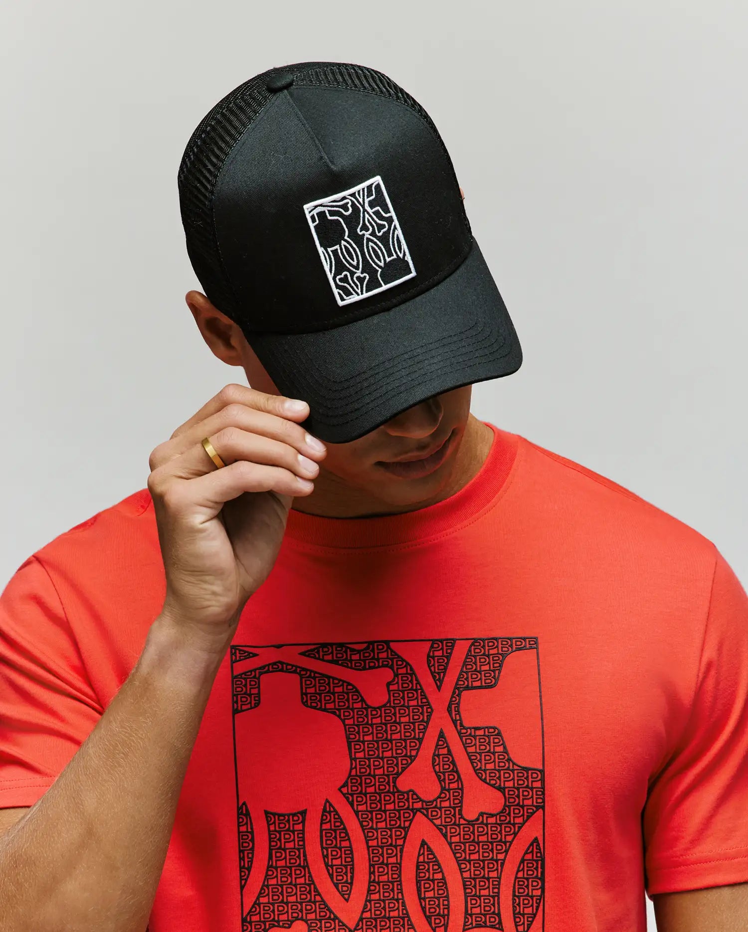 A person wears the Psycho Bunny MENS GLENN TRUCKER CAP - MO1500004, an adjustable black snapback with a white abstract pattern, paired with a red shirt featuring a matching black pattern. The background is plain and light-colored.