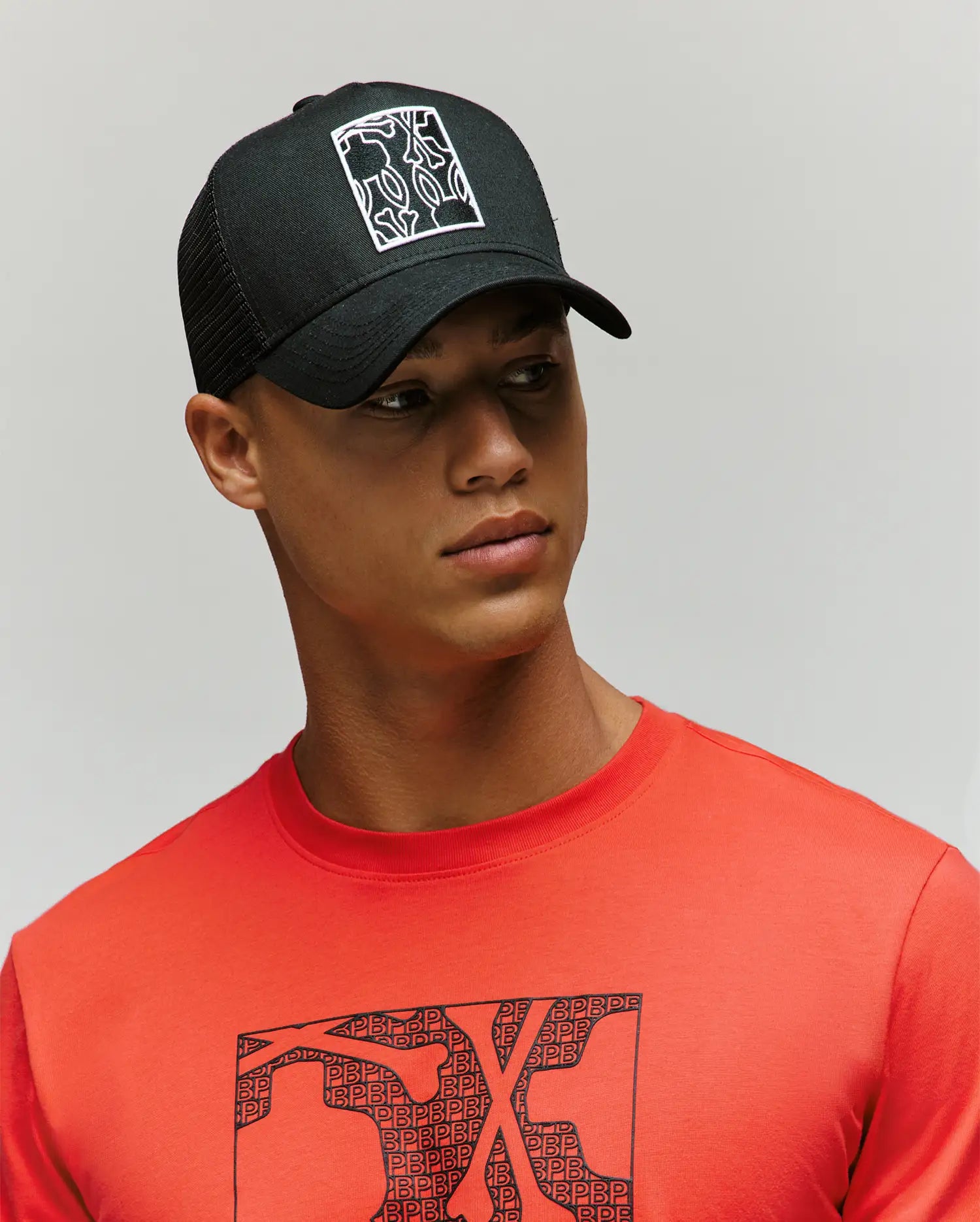 A young man wears the Psycho Bunny MENS GLENN TRUCKER CAP (MO1500004), a black snapback with a white abstract design, and a red t-shirt with a matching bunny graphic, looking left against a plain light background.