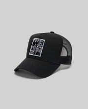 The Psycho Bunny MENS GLENN TRUCKER CAP (MO1500004) is a black trucker hat with mesh back, adjustable snapback, and a front square patch featuring a white-outlined abstract Bunny graphic, shown against a plain gray background.