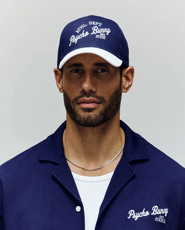 A bearded man wears a blue Psycho Bunny MENS MATTEO BASEBALL CAP (MO1500008) with an embroidered wordmark and a matching cotton twill shirt with white lettering, posing neutrally against a plain light background.
