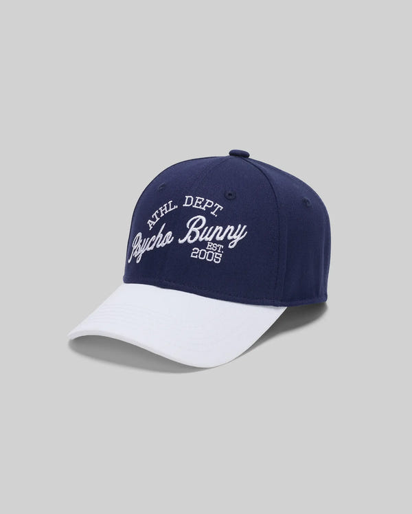 The Psycho Bunny MENS MATTEO BASEBALL CAP - MO1500008 is a navy cotton twill cap with a white brim and bold varsity-style "ATHL DEPT Psycho Bunny EST. 2005" embroidery on the front, shown against a plain grey background.