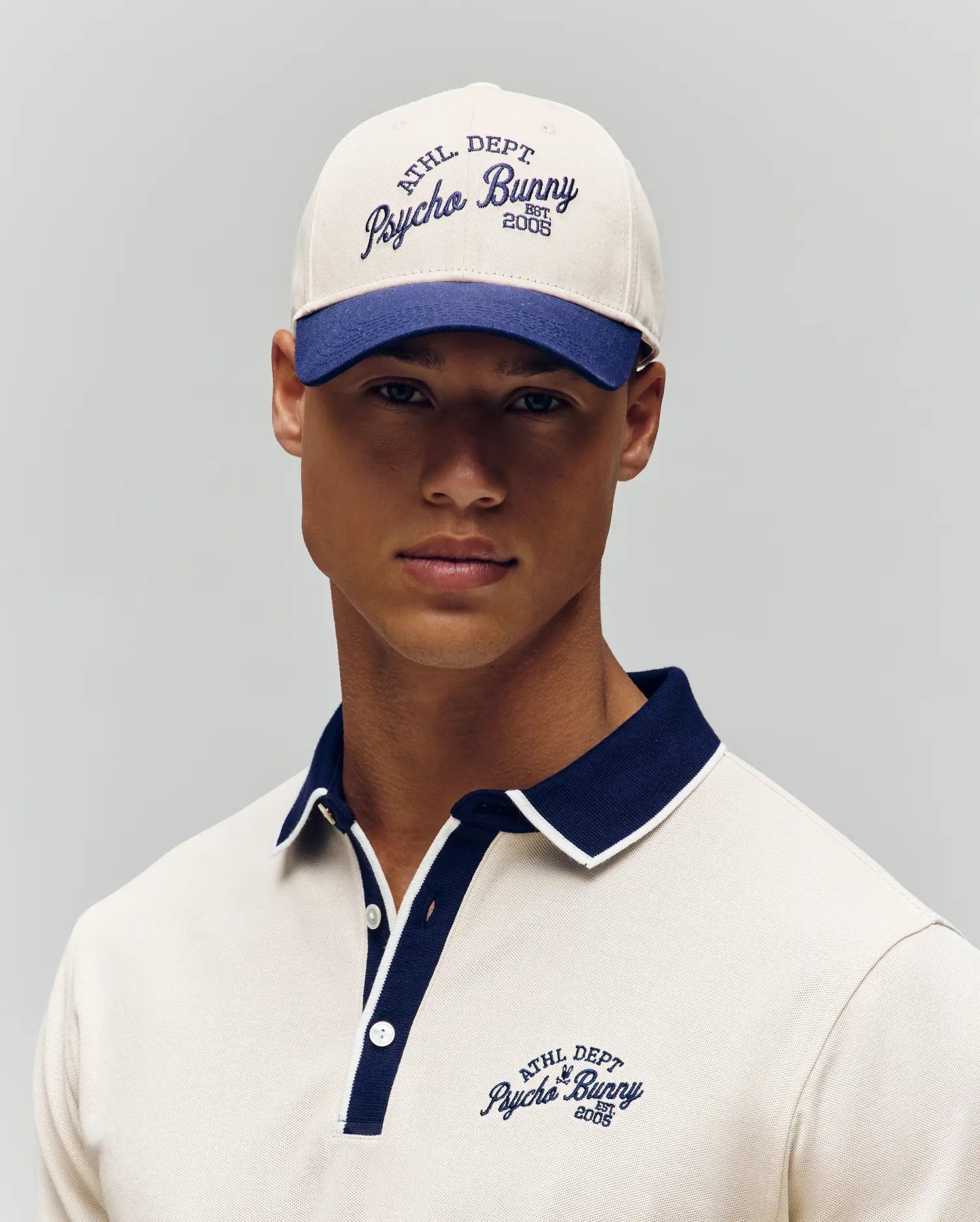 A person wearing the Psycho Bunny MENS MATTEO BASEBALL CAP - MO1500008 in a varsity cream and navy style, paired with a matching polo featuring embroidered 
