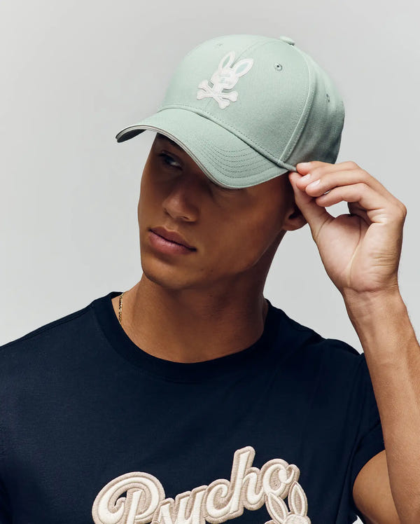 A person wearing the light green Psycho Bunny MENS DEREK BASEBALL CAP (MO1500009) with a white bunny logo and a black T-shirt featuring "Psycho" in bold beige script, looking off to the side while adjusting the cap.