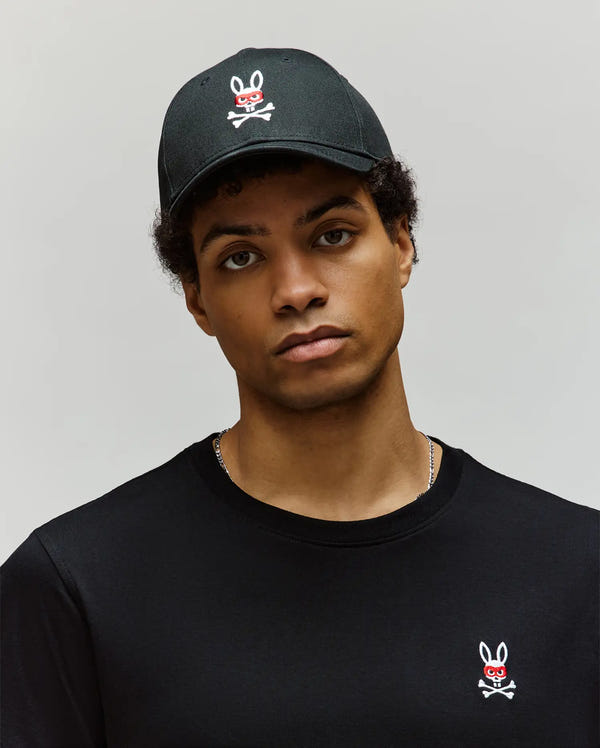 A young person in a black shirt and the Psycho Bunny MENS MISCHIEF BASEBALL CAP (MO1500015), both with the white Mischief Bunny skull and crossbones logo, stands against a plain light background, gazing neutrally at the camera.