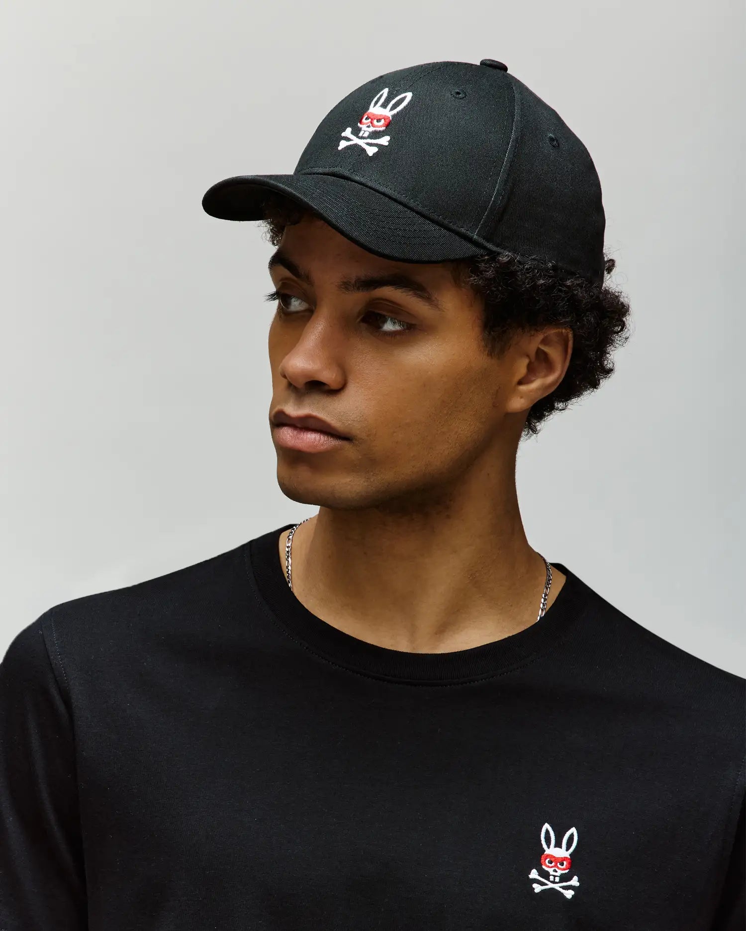A young man wears the Psycho Bunny MENS MISCHIEF BASEBALL CAP - MO1500015 and a black shirt, both 100% cotton with the signature Mischief Bunny logo, as he looks left against a plain light gray background.