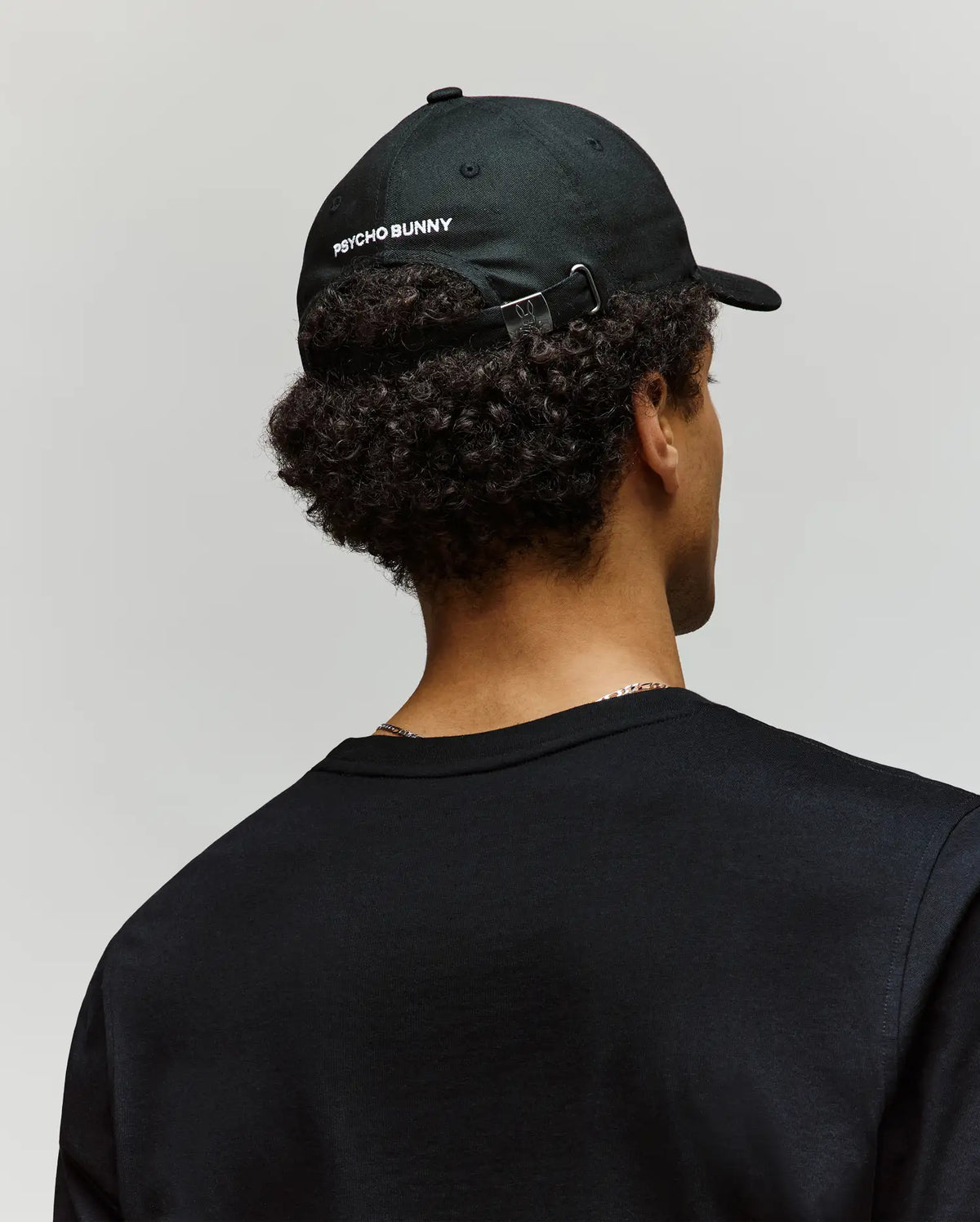 A person with curly hair, wearing a black Psycho Bunny MENS MISCHIEF BASEBALL CAP (MO1500015) made of 100% cotton and a black shirt, is shown from behind against a plain light background.