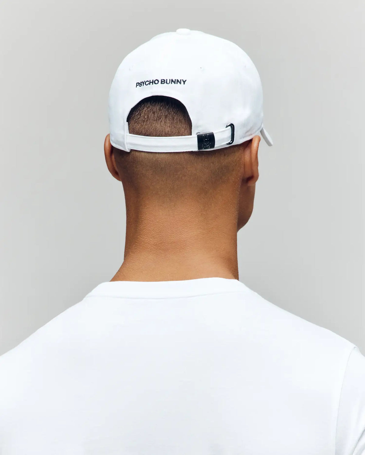 A person is shown from behind against a plain light background, wearing a white Psycho Bunny MENS MISCHIEF BASEBALL CAP (MO1500015) with "PSYCHO BUNNY" on the back, and a white 100% cotton shirt.