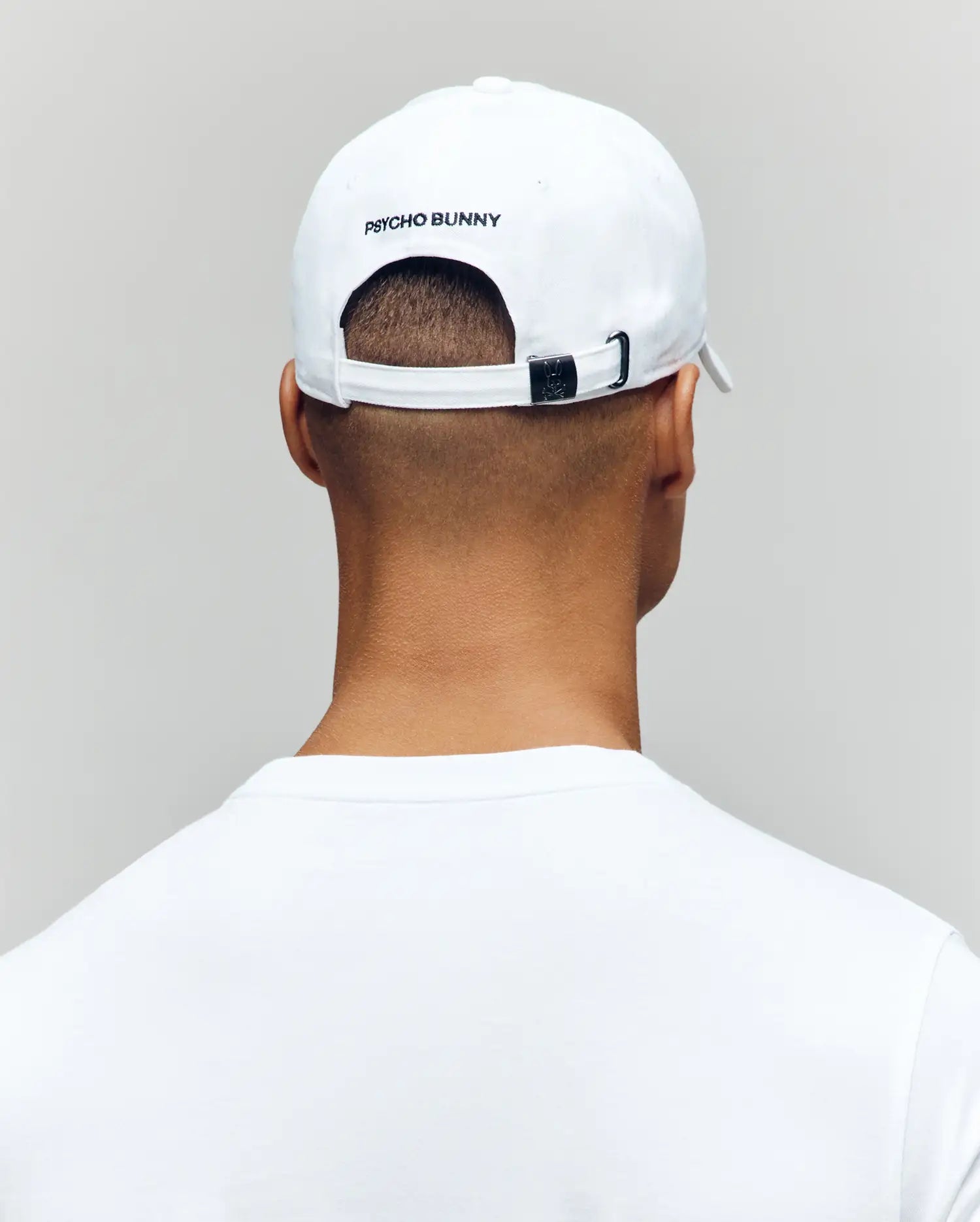 A person is shown from behind against a plain light background, wearing a white Psycho Bunny MENS MISCHIEF BASEBALL CAP (MO1500015) with "PSYCHO BUNNY" on the back, and a white 100% cotton shirt.