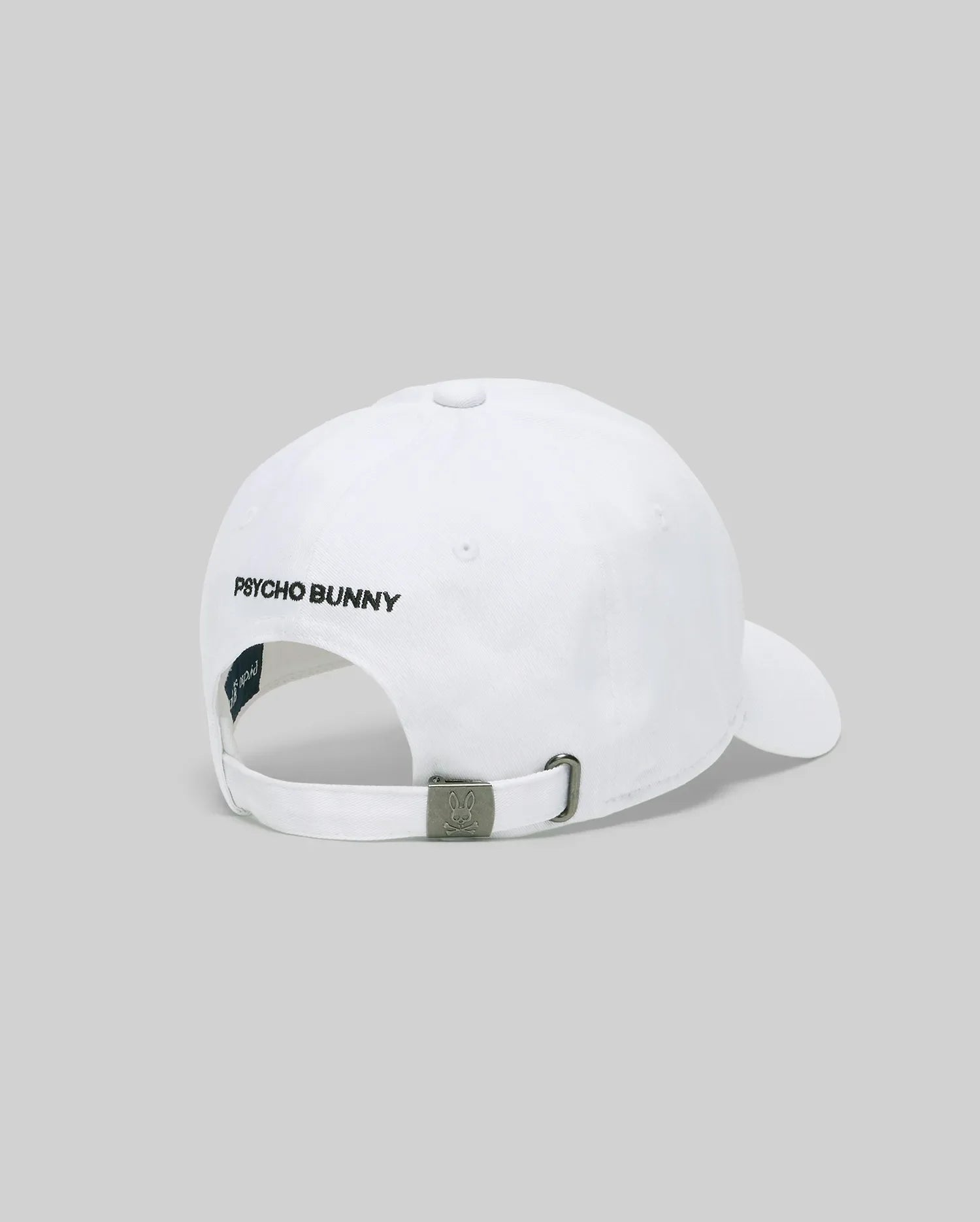 The Psycho Bunny MENS MISCHIEF BASEBALL CAP (MO1500015) is a white 100% cotton cap, shown from the back with "PSYCHO BUNNY" embroidered in black and an adjustable metal clasp strap against a plain light gray background.