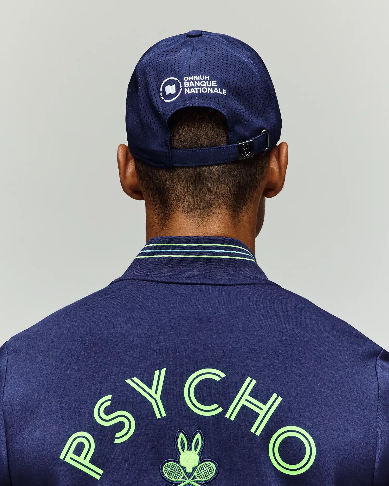 A person, seen from behind, wears a navy Psycho Bunny jacket with "PSYCHO" and a tennis bunny logo, plus the Psycho Bunny TENNIS BALL CREW MENS CAP FRENCH (MO1500025) featuring "OMNIUM BANQUE NATIONALE" on breathable mesh.