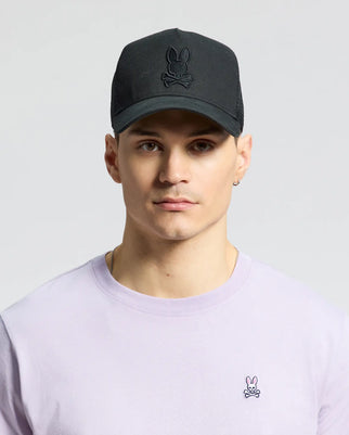 A person sports a Psycho Bunny MENS WILDER TRUCKER CAP (MO1500026) with an adjustable fit and breathable mesh, styled in black with a rabbit and crossbones emblem. They pair it with a light purple shirt featuring a small rabbit emblem, set against a plain gray backdrop.
