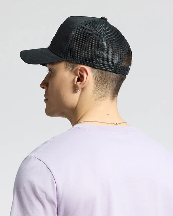 A person, seen from behind, wears a Psycho Bunny MENS WILDER TRUCKER CAP - MO1500026 in rugged black with an adjustable strap and a light purple t-shirt. With short hair and small hoop earrings, the individual stands against a plain, light-colored background.