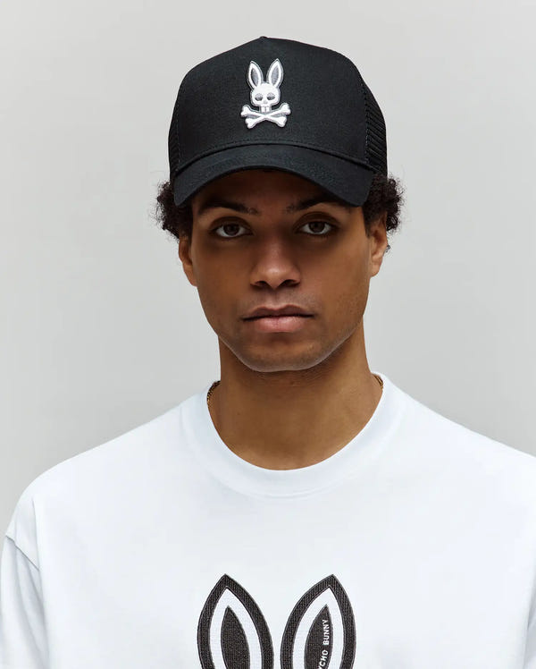 A person in a Psycho Bunny MENS RYDER TRUCKER CAP (MO1500030)—a black cotton twill snapback featuring a white skull and crossbones bunny logo—and a matching white shirt stands against a plain light gray background.