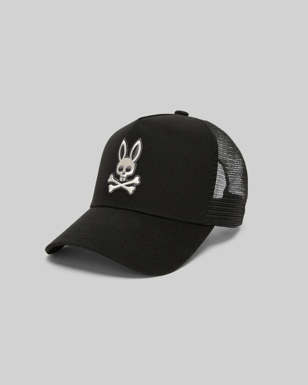 The Psycho Bunny MENS RYDER TRUCKER CAP (MO1500030) is a black mesh-back baseball cap in cotton twill, featuring a white embroidered bunny skull and crossbones on the front and an adjustable snapback, shown on a plain light gray background.