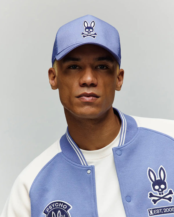 A person poses against a plain background wearing the Psycho Bunny MENS RYDER TRUCKER CAP - MO1500030 in light blue cotton twill, featuring a cartoon bunny with crossbones logo and an adjustable snapback for a perfect fit.